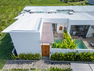 Aerial view of a white villa, its timber gate and a planted courtyard with pool beside a green field.