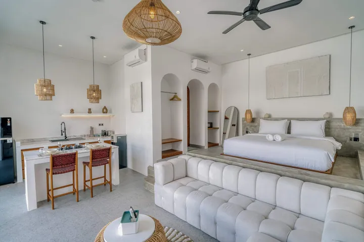 Open-plan bedroom and lounge with white bed, sectional sofa and woven pendant lights.