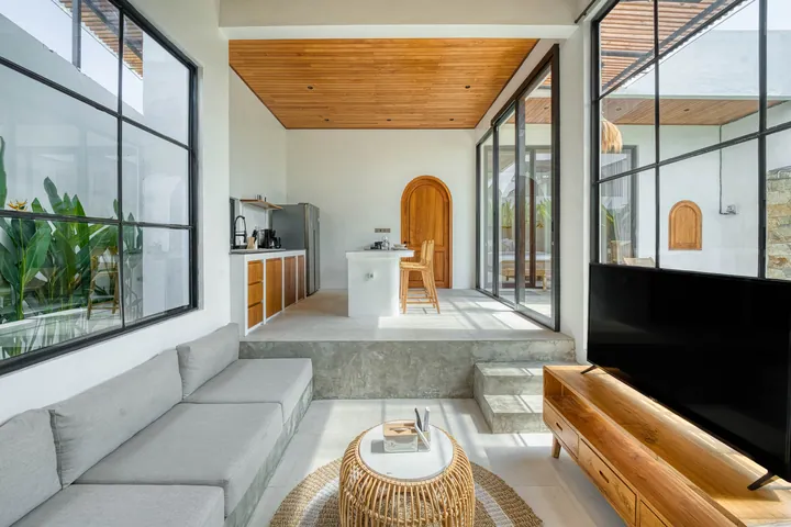 Pale seating and a television beneath a timber ceiling, beside floor-to-ceiling windows.