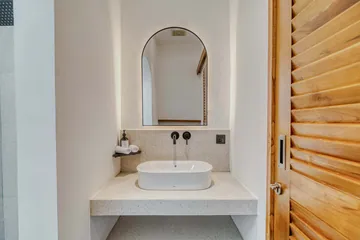 Pale stone vanity with a vessel basin and arched mirror beside a louvred timber door.