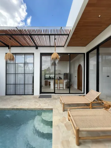 Courtyard loungers and pool steps in front of black-framed glass rooms beneath a slatted pergola.
