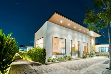 Illuminated white villa exterior at night with tall windows and planting along a paved lane.