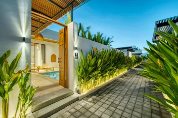 Paved lane between a planted border and a white villa wall, with a glimpse of a courtyard pool.
