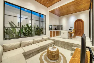 Living room with a sectional sofa and woven coffee table beneath a timber ceiling, with the kitchen beyond.