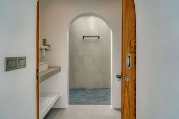 Walk-in shower beside a toilet and wood slatted door in a pale stone bathroom.