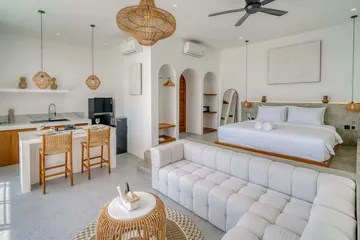 Open-plan bedroom and lounge with white bed, sectional and kitchen counter.