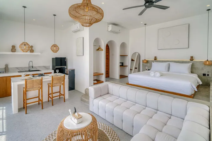 Open-plan bedroom and lounge with white bed, sectional and kitchen counter.