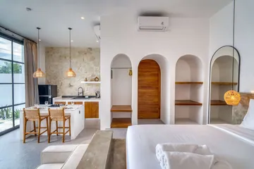 Kitchen counter and bed seen across a bright room with arched wooden doorway and shelving.