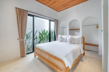 White bed on a wood frame beside a curtained window and potted greenery.