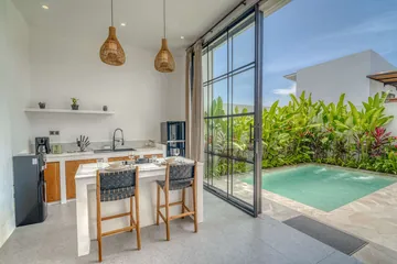 Timber kitchen cabinetry and a dining island beside open glass doors and a courtyard pool.