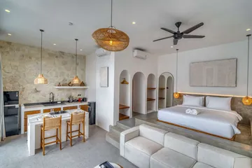 Open-plan room with a low bed, pale sectional sofa, stone kitchen wall and woven pendant lights.