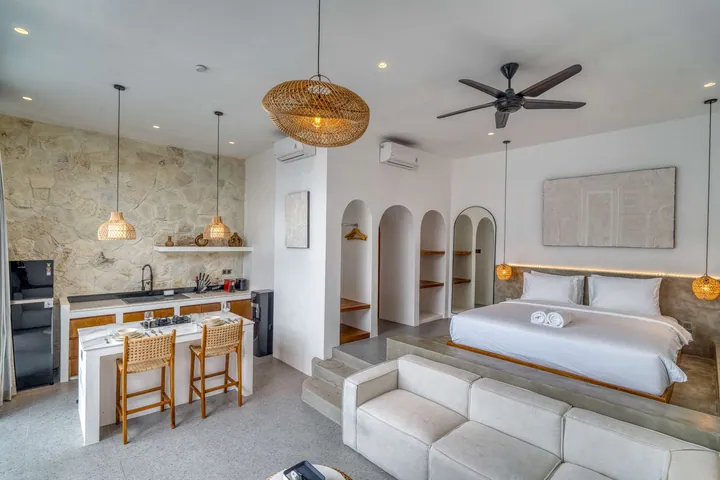 Open-plan room with a low bed, pale sectional sofa, stone kitchen wall and woven pendant lights.