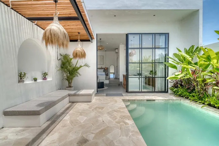 A courtyard with a built-in bench, woven lamps, pool and tall tropical leaves.