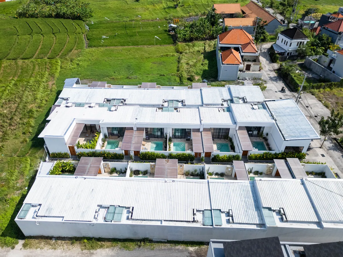 Aerial view of a row of white villas with courtyard pools and timber pergolas beside green fields and neighbouring red roofs.