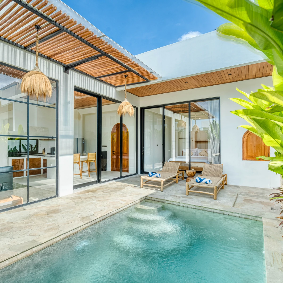 A courtyard pool between glass-walled rooms, with timber roof slats and broad green leaves.